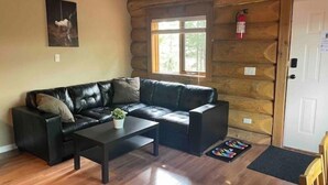 A log cabin interior with a black leather sofa, a small coffee table with a plant, wooden floors, a window with a view, and a fire extinguisher on the wall. - Dogs love our cabin in Lac Le Jeune close to lake (Logan Lake)