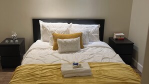 A neatly made bed with white and yellow bedding, two bedside tables with small items, and a plain wall with a ceiling light.