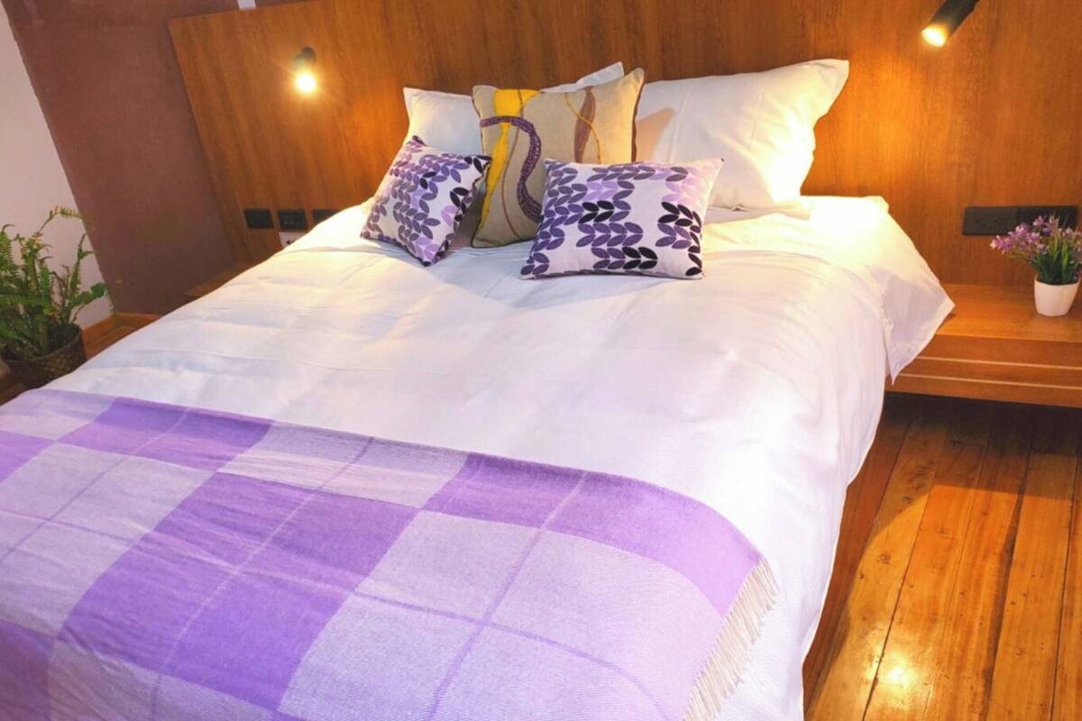 Un letto rifatto con cura, coperta a quadretti viola, numerosi cuscini e una testiera in legno.