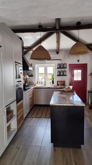 A kitchen with a large island, wooden beams, hanging light fixtures, and a red door. - Casa Vó Briata (Mértola)