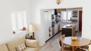 A modern kitchen with white cabinetry, a dining area with a wooden table and wicker chairs, and a cozy living room with a beige sofa. - Casa Vó Briata (Mértola)