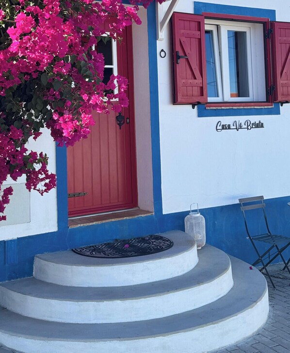 A building with a red door, a window with red shutters, and a sign that reads 'Casa Vo' Briola'. - Casa Vó Briata (Mértola)