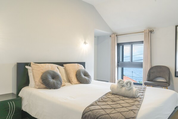 Habitación de hotel con cama, una silla, ventana con cortinas y vista al mar.
