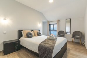 A modern bedroom with a bed, bedside tables, a window with curtains, and a view of the sea.