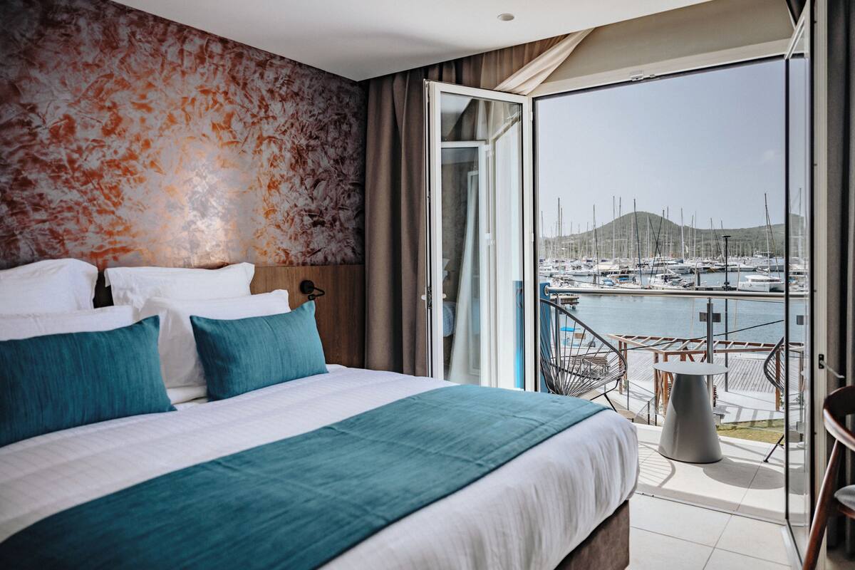 A hotel room with a bed, a view of a marina, and a balcony with outdoor seating.