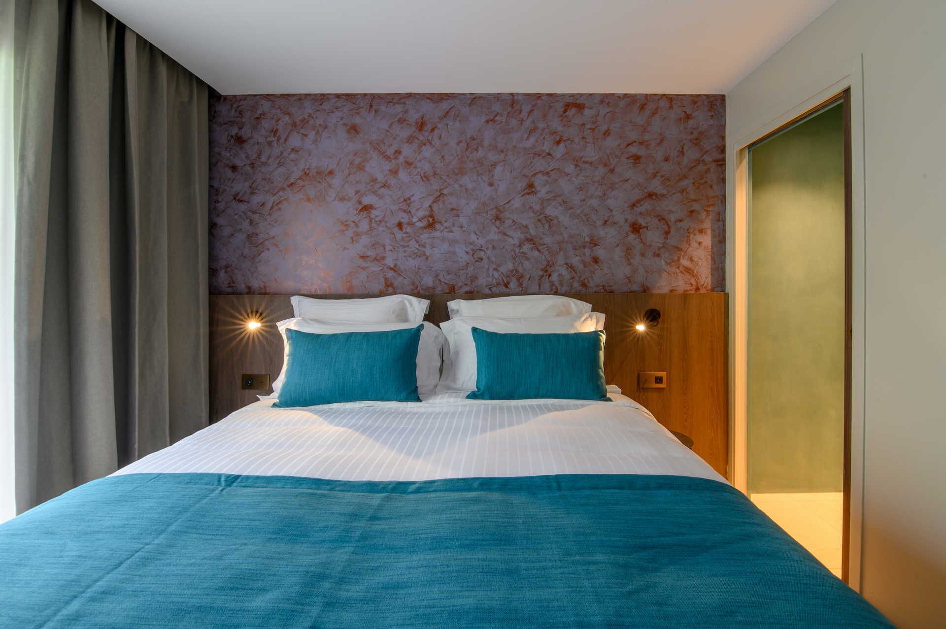 Camera d'albergo con un letto grande, biancheria da letto color petrolio e carta da parati fantasia.
