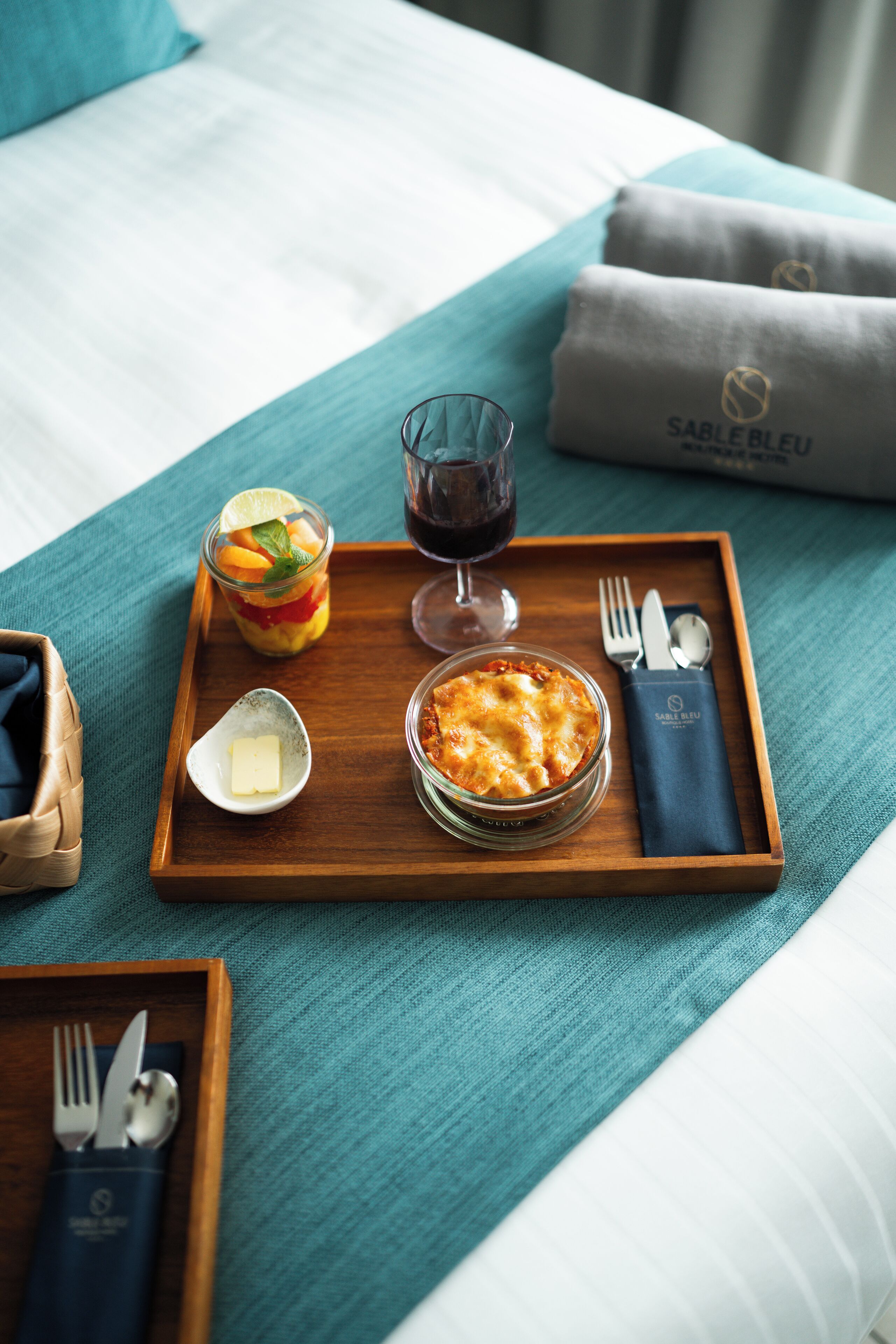 A neatly arranged breakfast tray on a bed with a teal bedspread, including a glass of red wine, a fruit salad, a small bowl of butter, and a dish of baked potatoes.