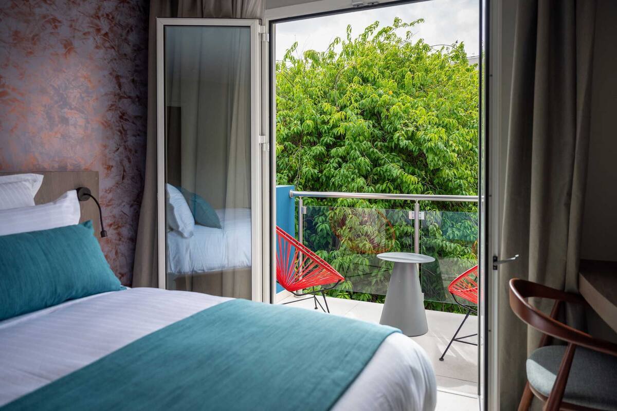 A hotel room with a bed, a small table, and a chair. There is a balcony with a view of trees.