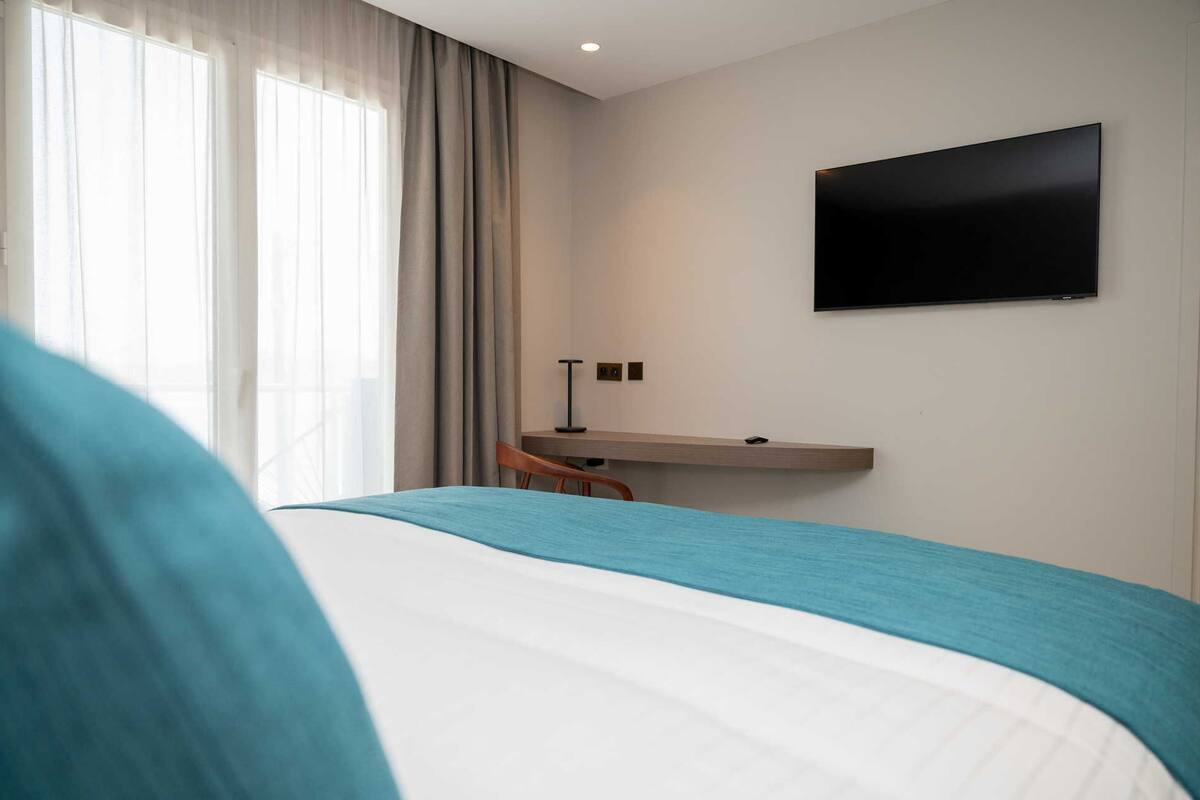 A hotel room with a bed, a desk, a chair, a TV mounted on the wall, and a window with curtains.