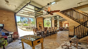 Un vaste espace intérieur comprenant une table de baby-foot, un sol en pierre, un escalier en bois et une vue sur l’extérieur.