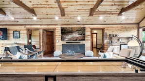 Amplia sala de estar con techo de madera, una televisión de pantalla plana grande sobre la chimenea, y una zona de bar con un reloj y taburetes.