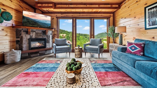 Interior acogedor de una cabaña con chimenea de piedra, dos sillones azules, un sofá azul y un ventanal con vistas al exterior.