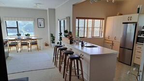 A modern kitchen with a central island, bar stools, a dining area with a table and chairs, and a refrigerator. - Comfort and the best seat in the west to view the Tasman Sea (Baylys Beach)