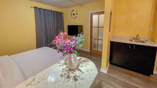 Une pièce comprenant un lit, une petite table avec un vase de fleurs, un coin cuisine et une porte dotée de panneaux en verre dépoli.