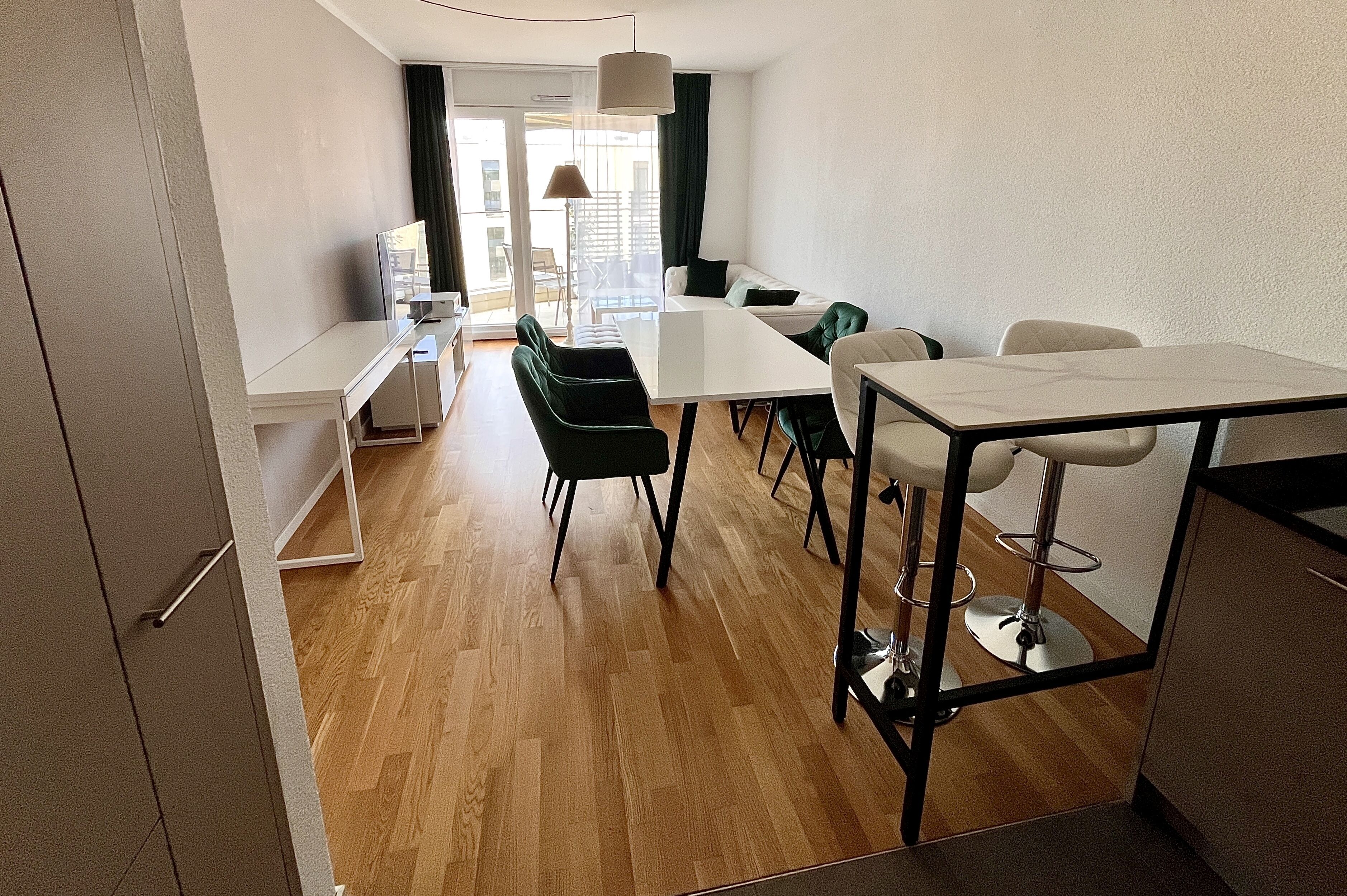 A modern, well-lit room with a wooden floor, a white dining table, black chairs, a white sofa, and a TV on a stand.