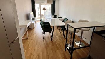 A modern, well-lit room with a wooden floor, a white dining table, black chairs, a white sofa, and a TV on a stand.