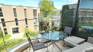A balcony with a wicker sofa, a small table, and a chair. There is a view of buildings and greenery in the background.