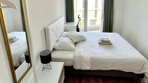A hotel room with a large bed, a bedside table, a chair, a mirror, and a view of the outside.