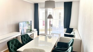A modern hotel room with a large dining table, green upholstered chairs, a white sofa, a flat-screen TV, and a view of the outdoors.