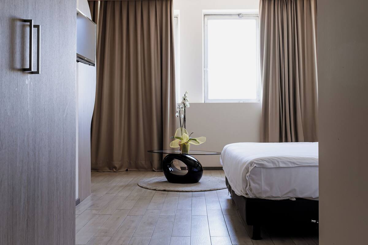 Ein modernes Schlafzimmer mit einem Bett, einem runden Tisch mit einer Vase und einem Fenster mit Vorhängen.