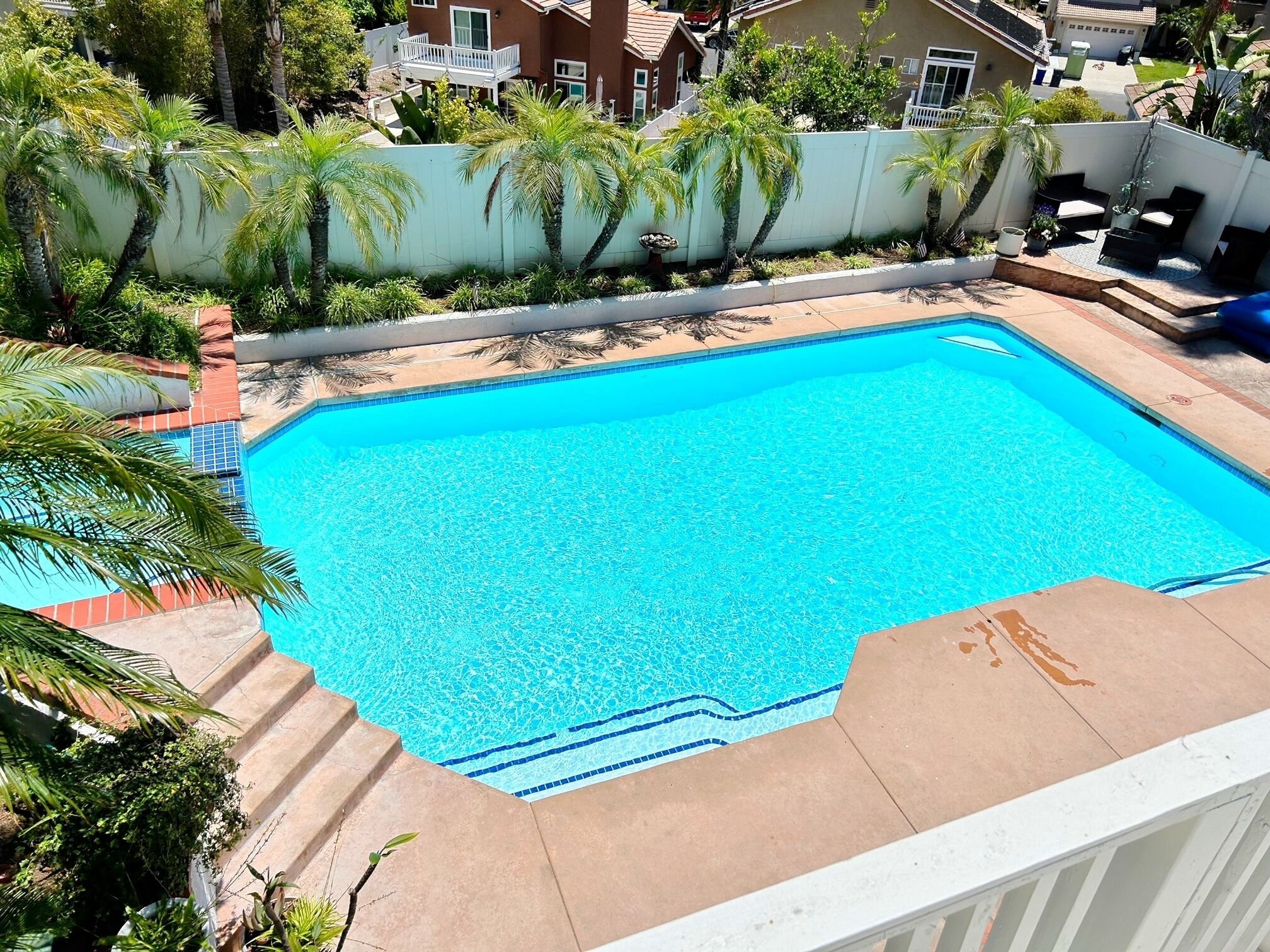 A rectangular swimming pool with clear blue water, surrounded by a white fence and palm trees.
