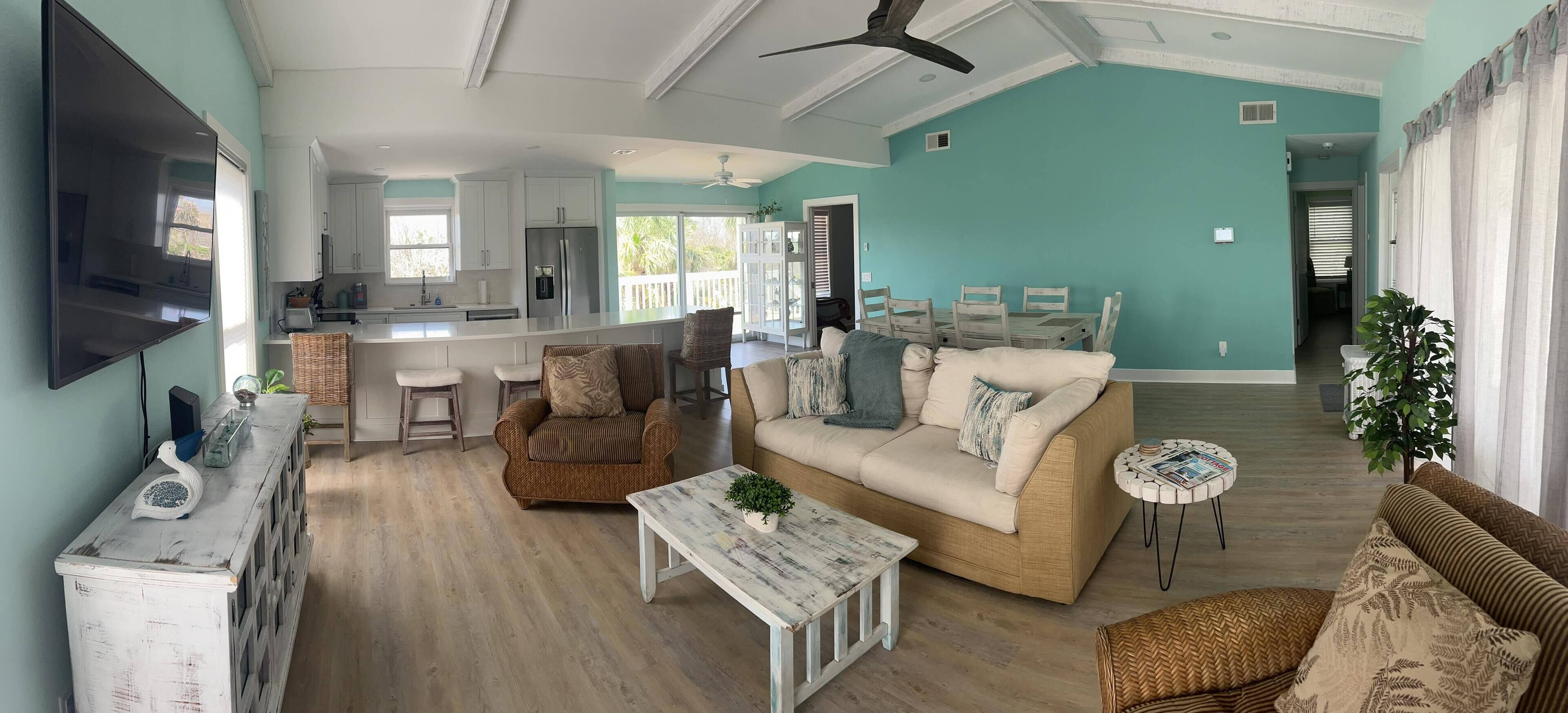 A spacious living area with a sofa, coffee table, and a dining table. A kitchen with white cabinets and a refrigerator is visible in the background.