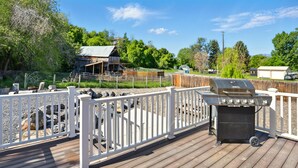 A wooden deck with a barbecue grill, white railings, and a fenced area with a barn and trees in the background.