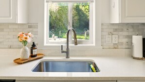 A kitchen with a stainless steel sink, a window with a view of greenery, white cabinets, and a wooden tray with a vase and a bottle.