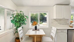 A kitchen with a white countertop, a dining table with two chairs, a potted plant, and a window with a view of greenery.