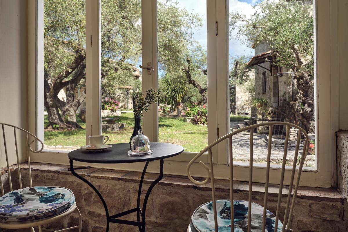 A room with a view of a garden through large windows, a small table with a vase, and two chairs.