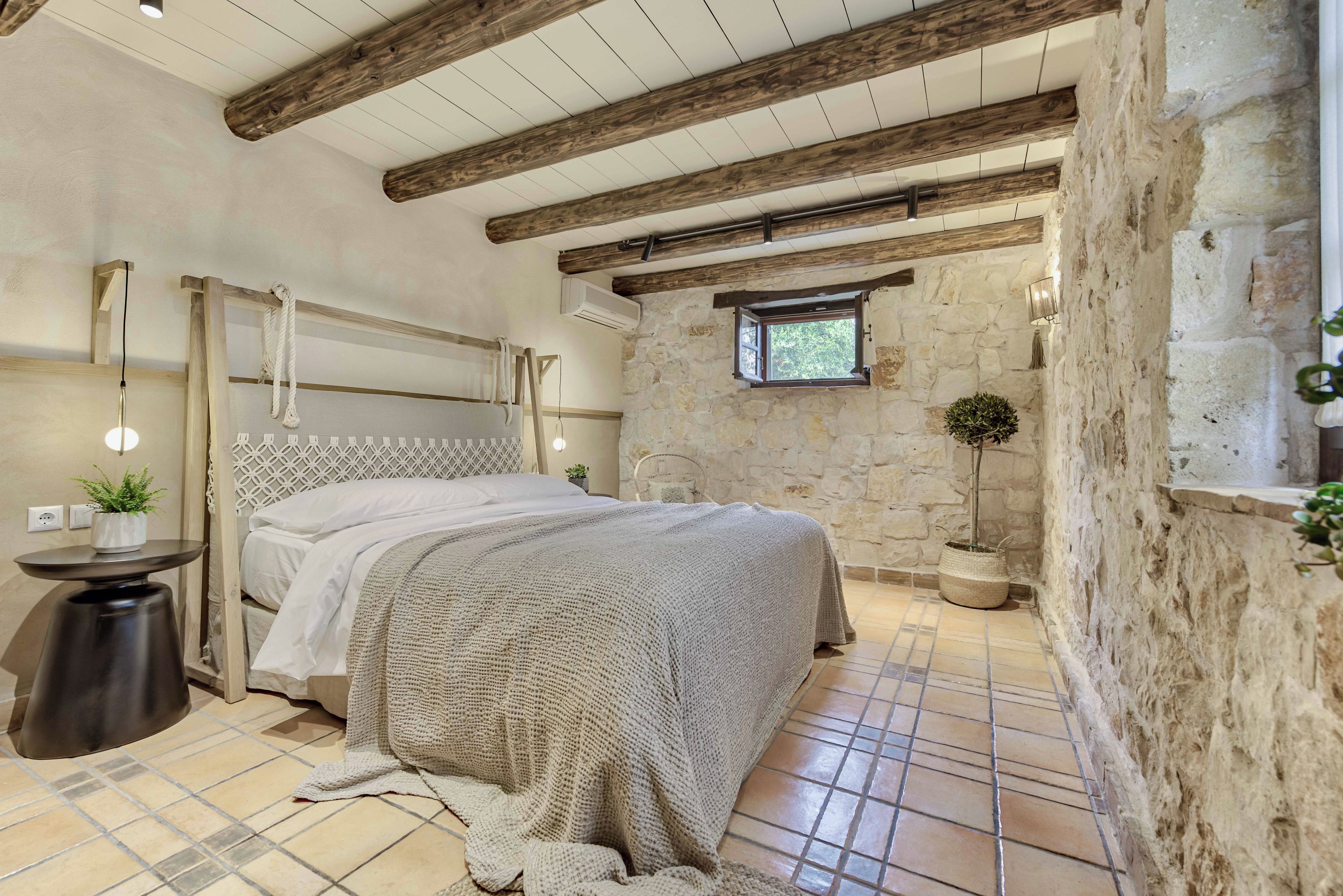 Un dormitorio con pared de piedra, techo de madera, cama con manta tejida, una ventana pequeña y una planta en maceta.