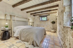 Ein Schlafzimmer mit Steinsandsteinwand, Holzbalkendecke, einem Bett mit Strickdecke, einem kleinen Fenster und einem Topfpflanzen.