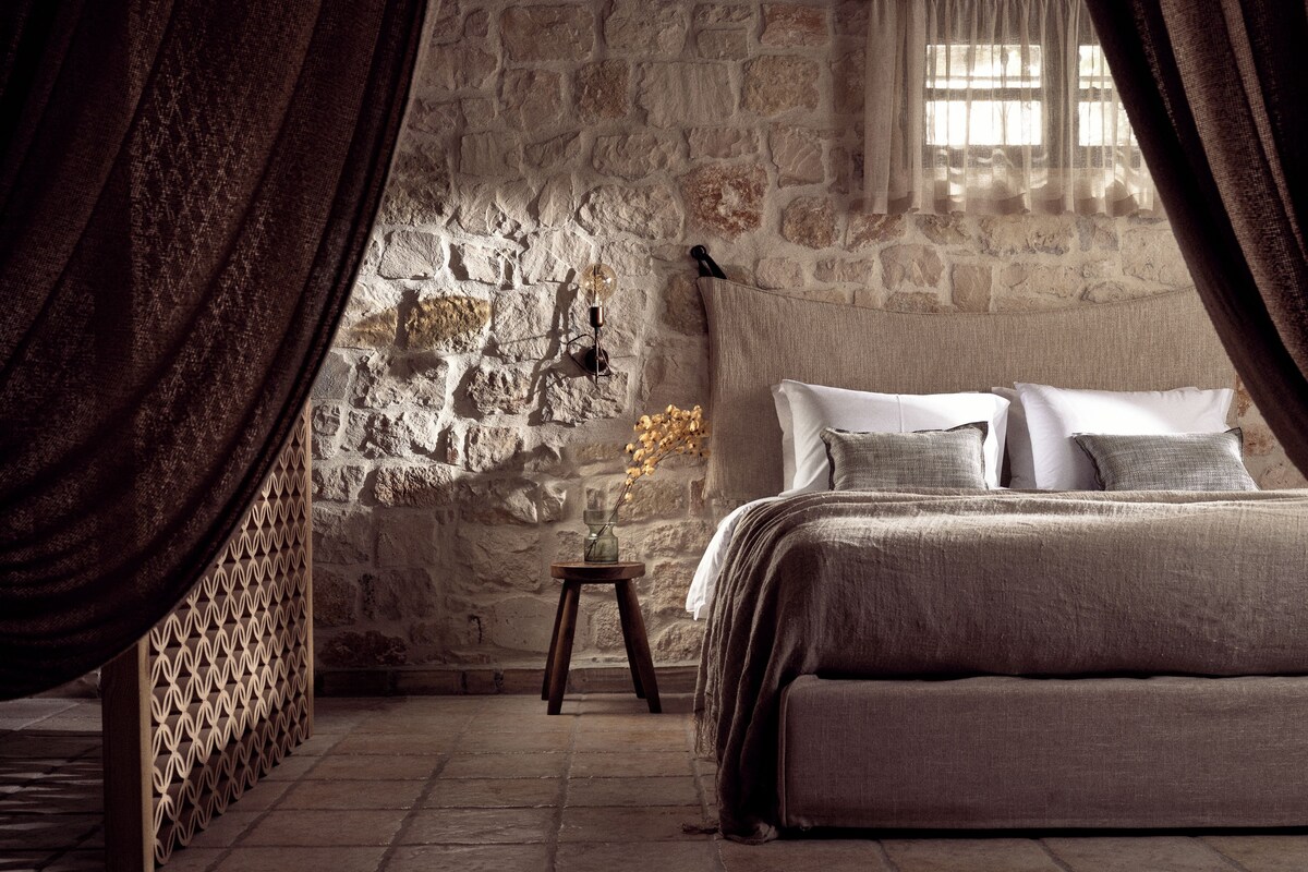 Un dormitorio con pared de piedra, una cama con muchas almohadas, una mesita con un florero y una ventana con cortinas.