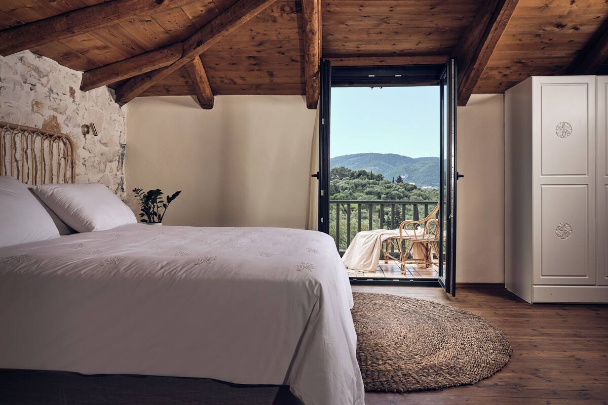 A bedroom with a wooden ceiling, a bed with white linens, a view of greenery and hills through an open door, and a wicker chair outside.