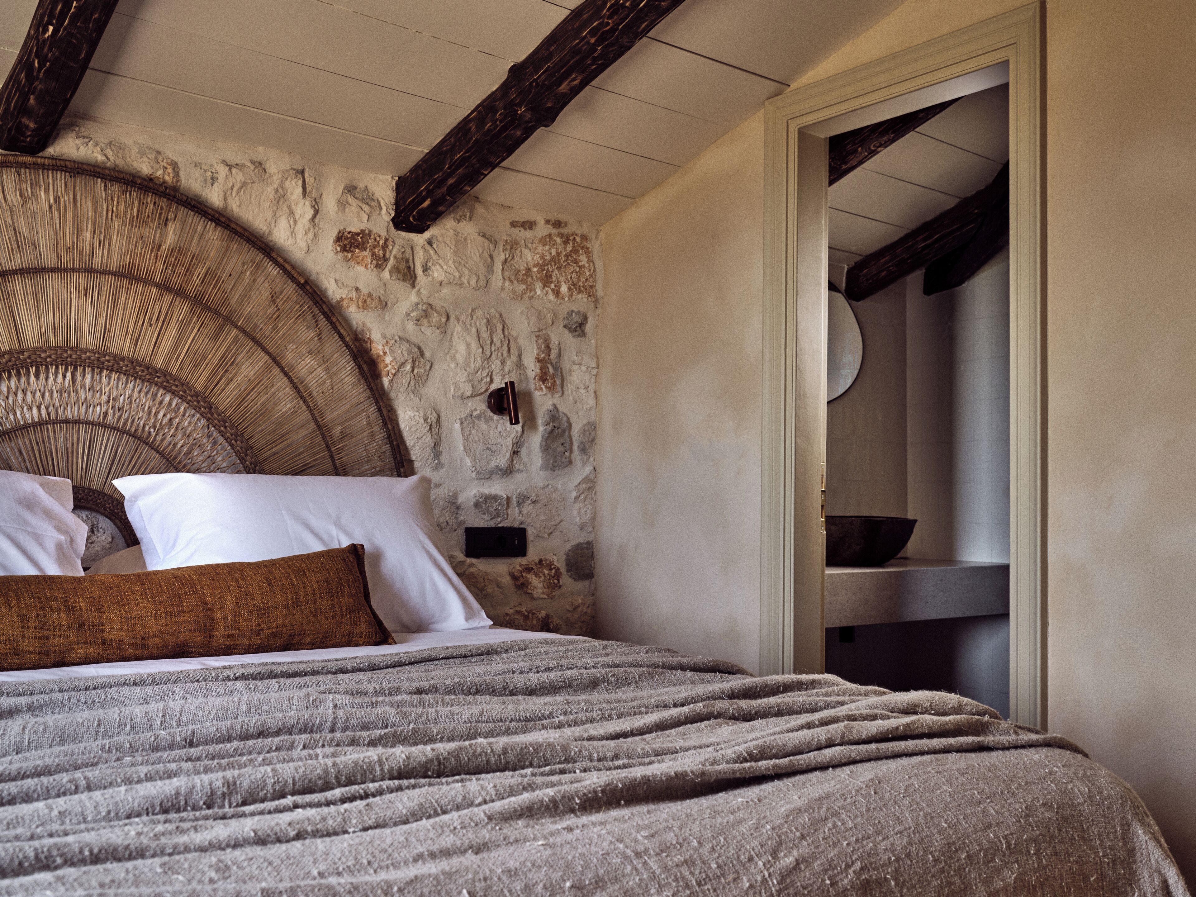 Une chambre avec un mur en pierre, une tête de lit en bois et un lit recouvert d’une couverture et de coussins gris.