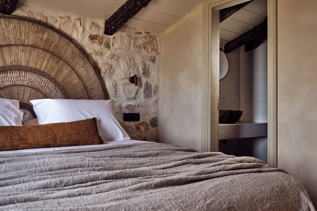 A bedroom with a stone wall, a wicker headboard, a bed with a grey blanket, and a wooden door.