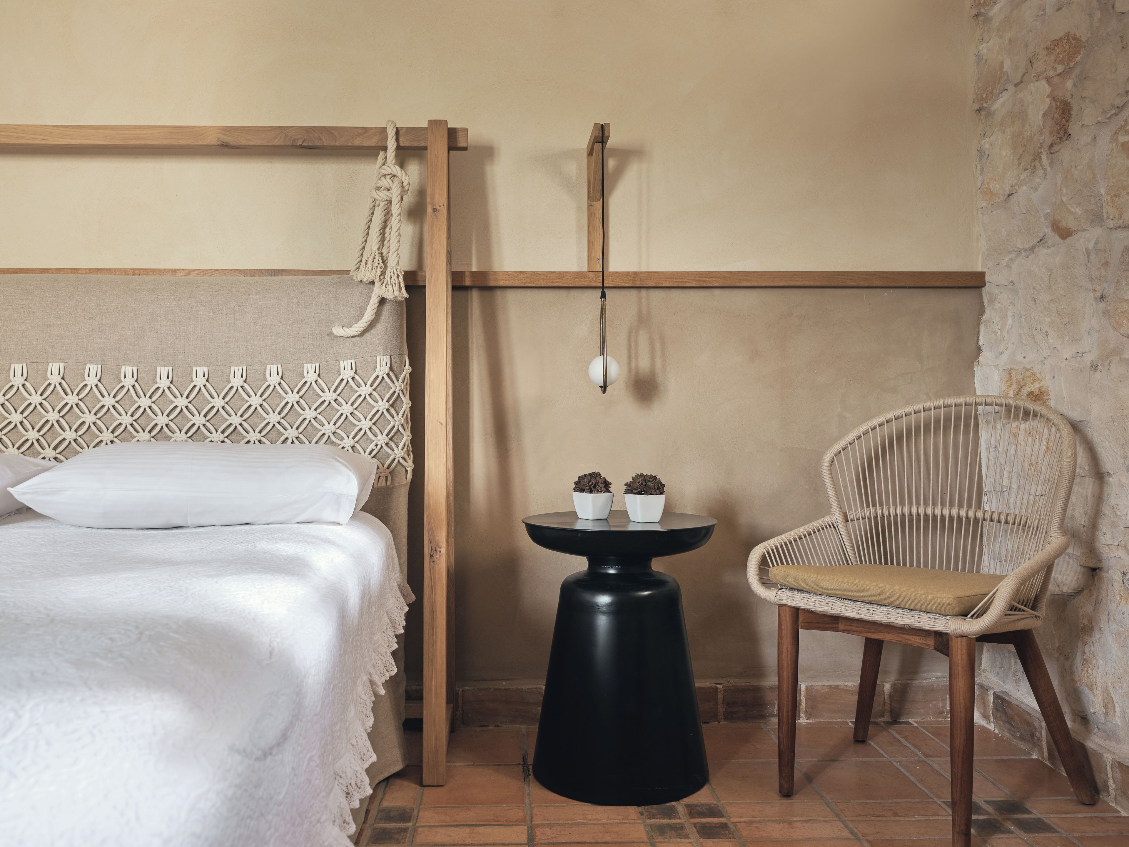 A room with a bed, a small round table with two potted plants, a wicker chair, and a macrame wall hanging.