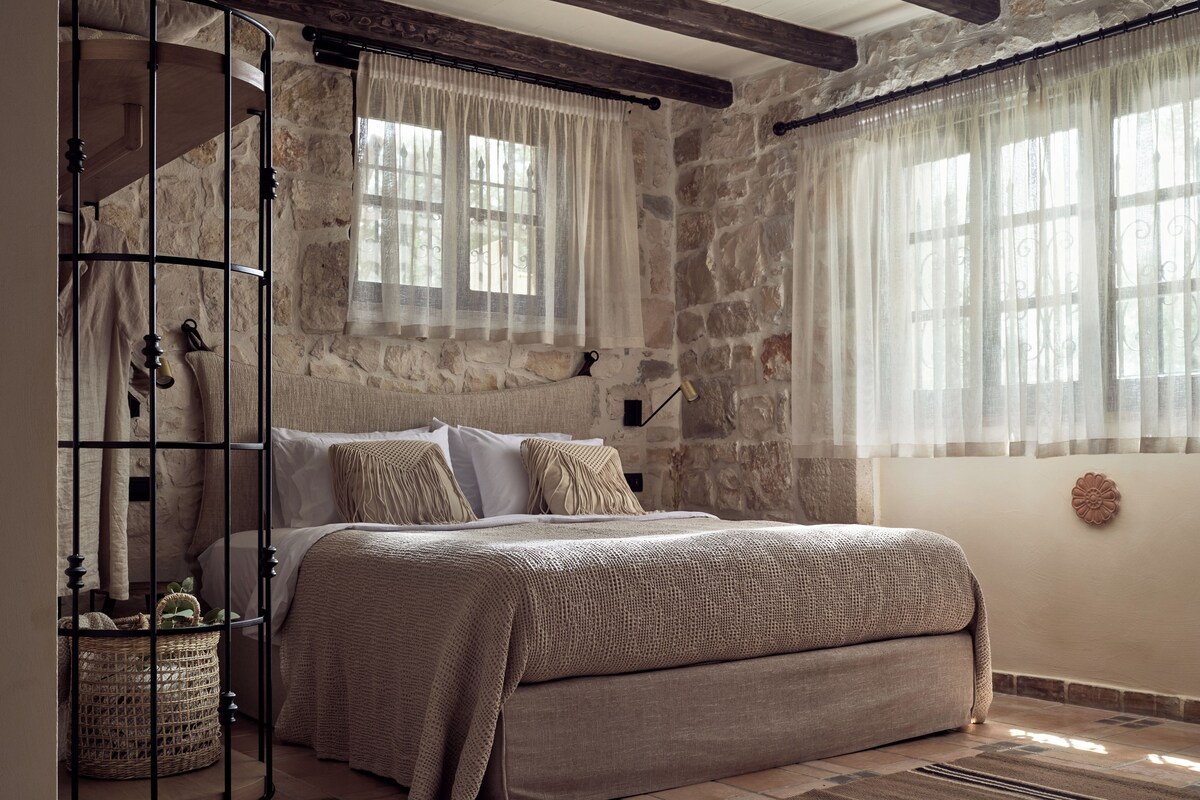 A bedroom with a stone wall, a bed with a beige blanket, a wicker basket, and a wooden headboard.