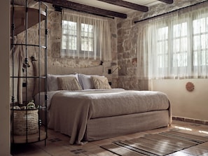 Une chambre avec un mur en pierre, un lit recouvert d’une couverture beige, un panier en osier et une tête de lit en bois.