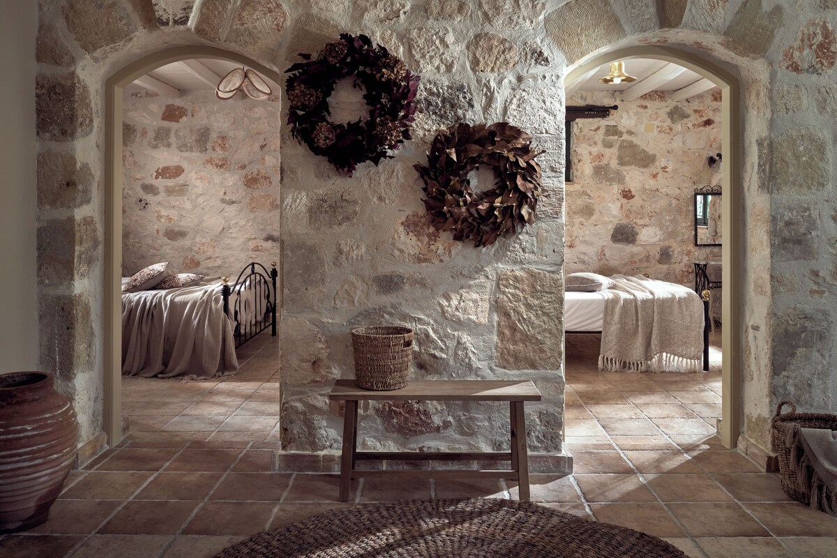 A rustic stone-walled room with two beds, a woven bench, and two large wreaths on the wall.