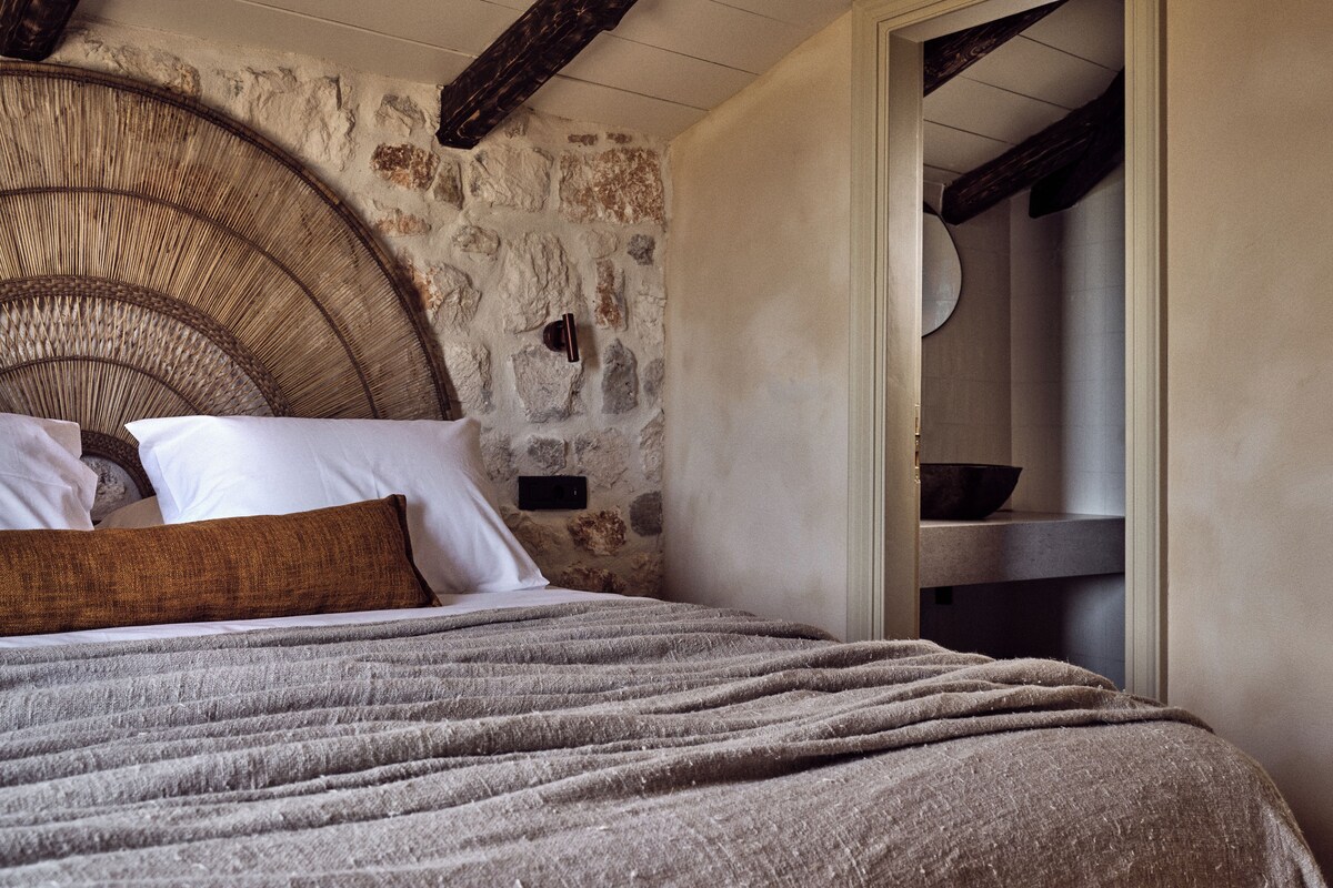 Un dormitorio con pared de piedra, cabecero de mimbre, cama con manta gris y puerta de madera.