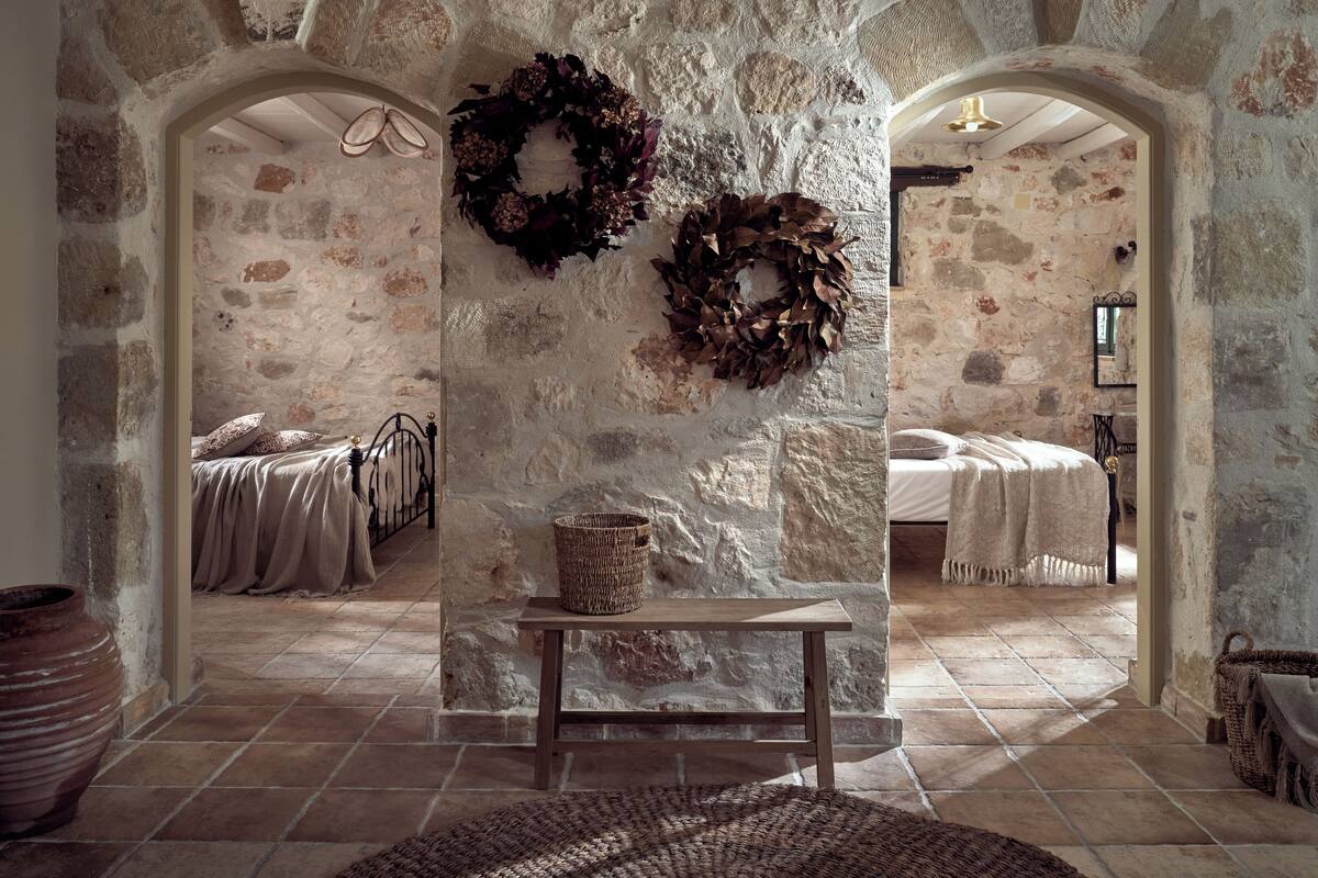 A rustic stone-walled room with two beds, a woven bench, and two large wreaths on the wall.
