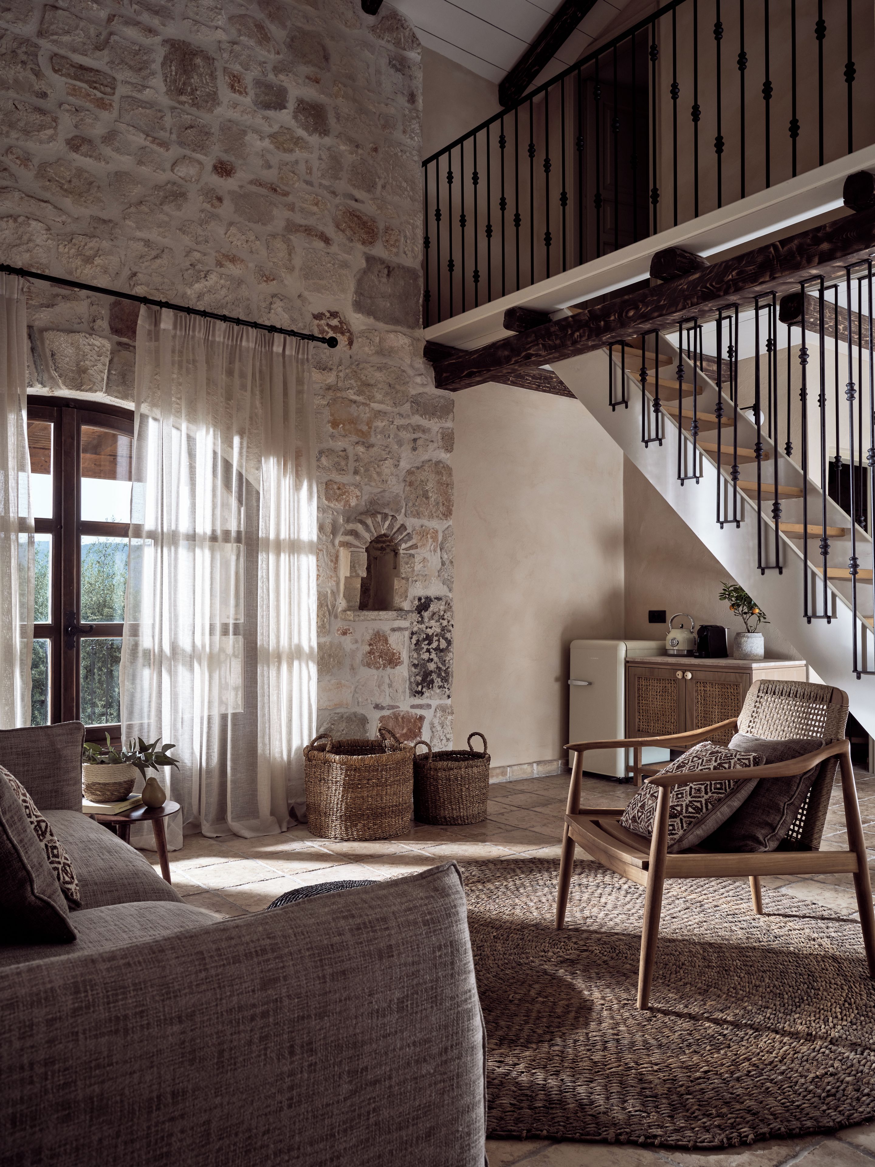 Un interior rústico con pared de piedra, una escalera de madera, un sofá y un sillón de mimbre.