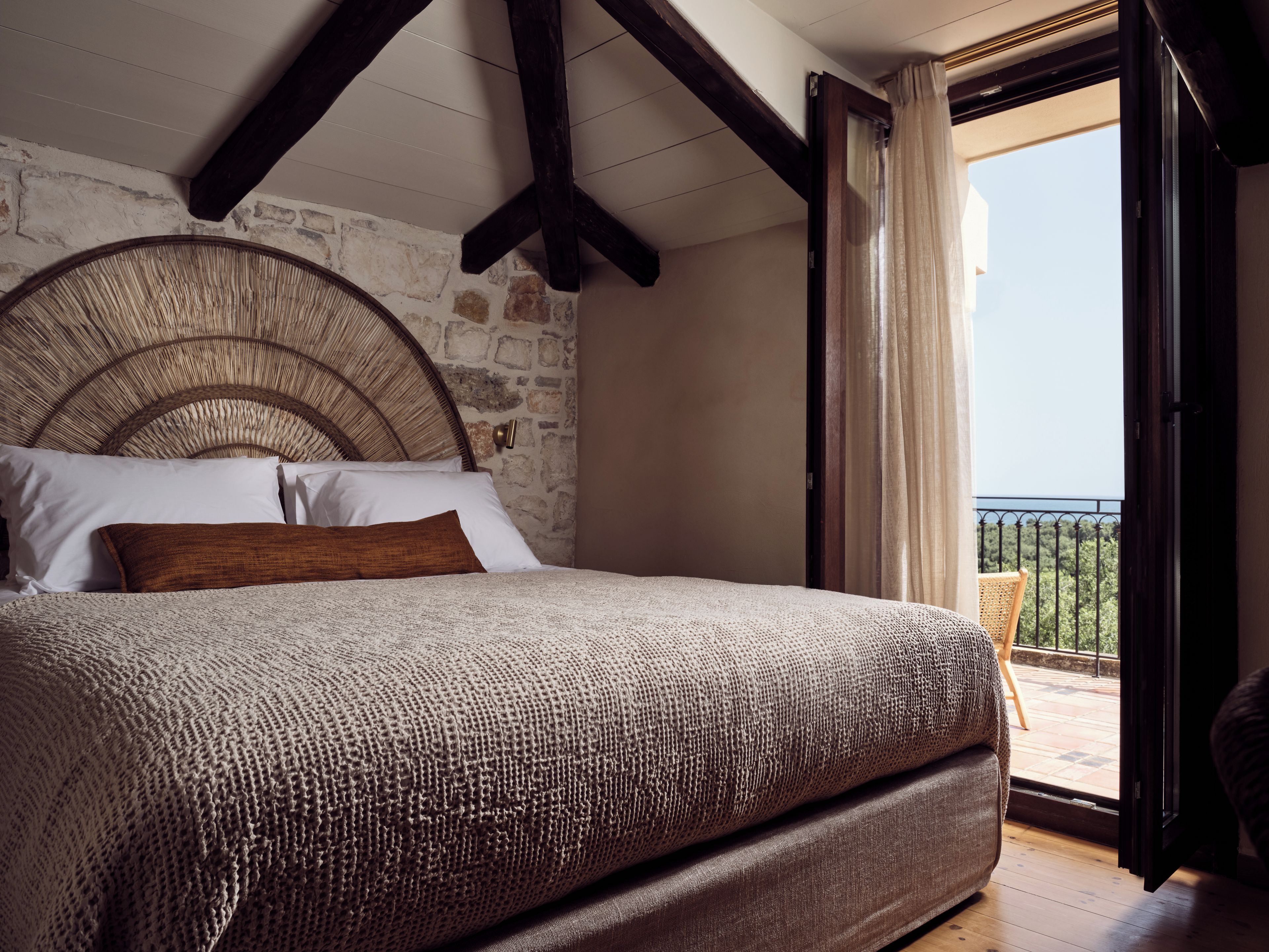 A bedroom with a stone wall, a large bed with a woven headboard, and a balcony with a view.