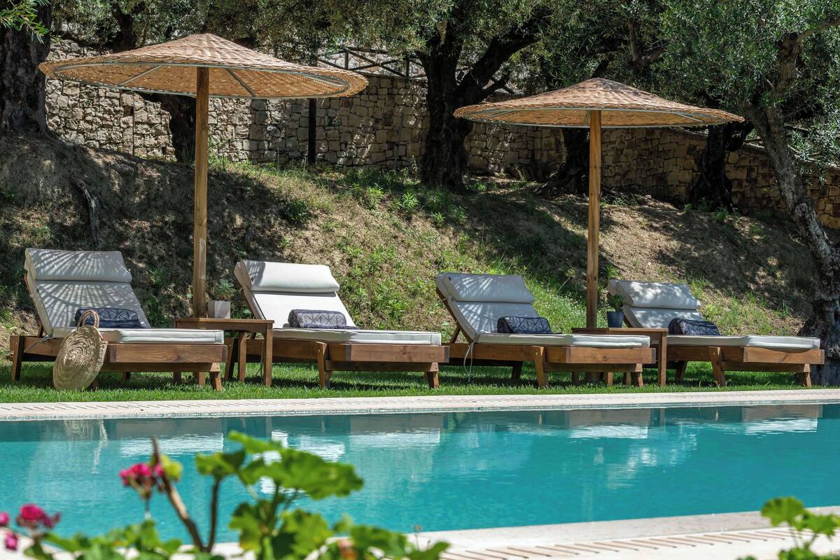 A poolside area with lounge chairs and umbrellas, surrounded by stone walls and greenery.