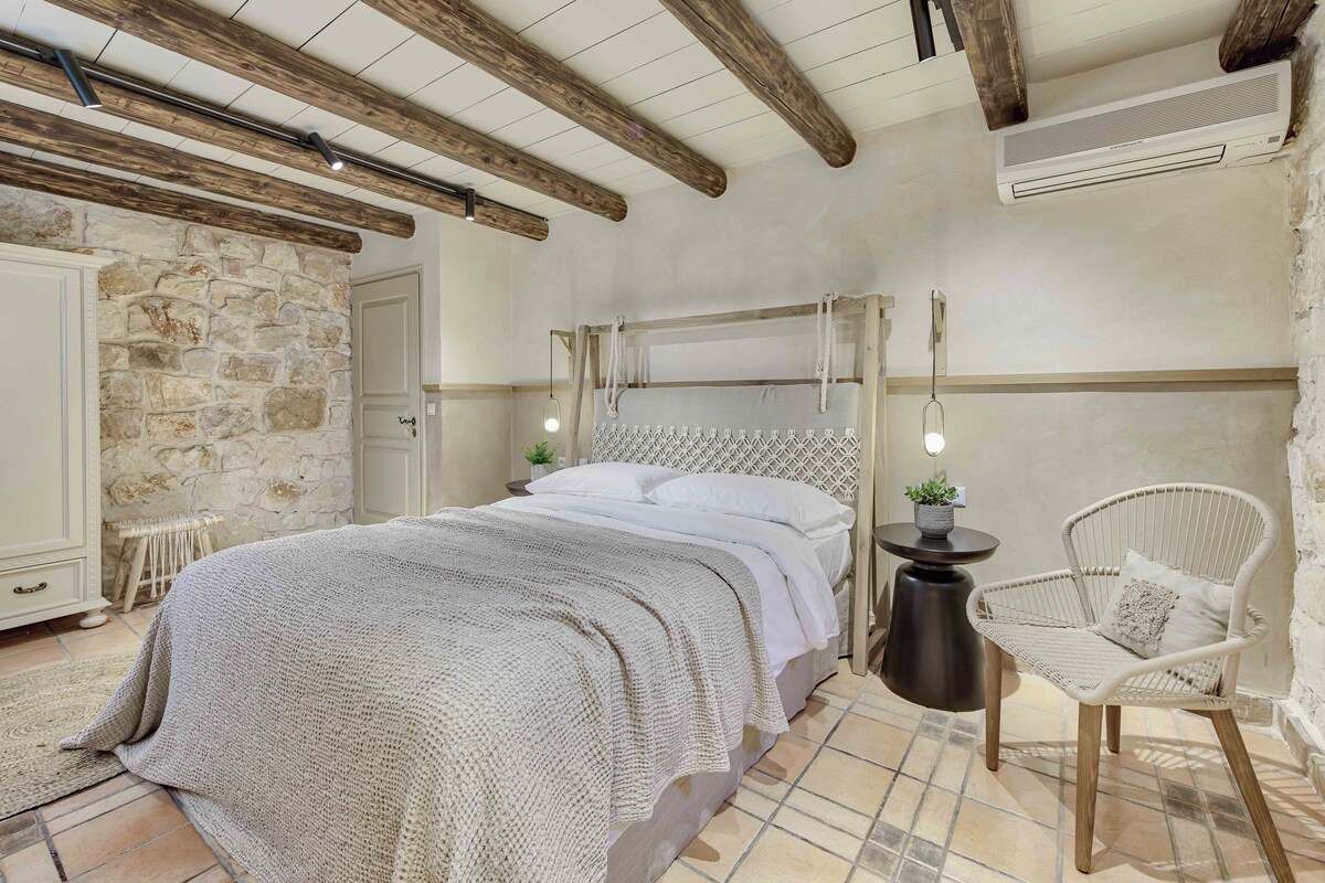 A bedroom with a stone wall, a bed with a knitted blanket, a wicker chair, and a small table with a plant.
