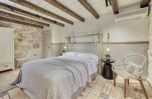 A bedroom with a stone wall, a bed with a knitted blanket, a wicker chair, and a small table with a plant.