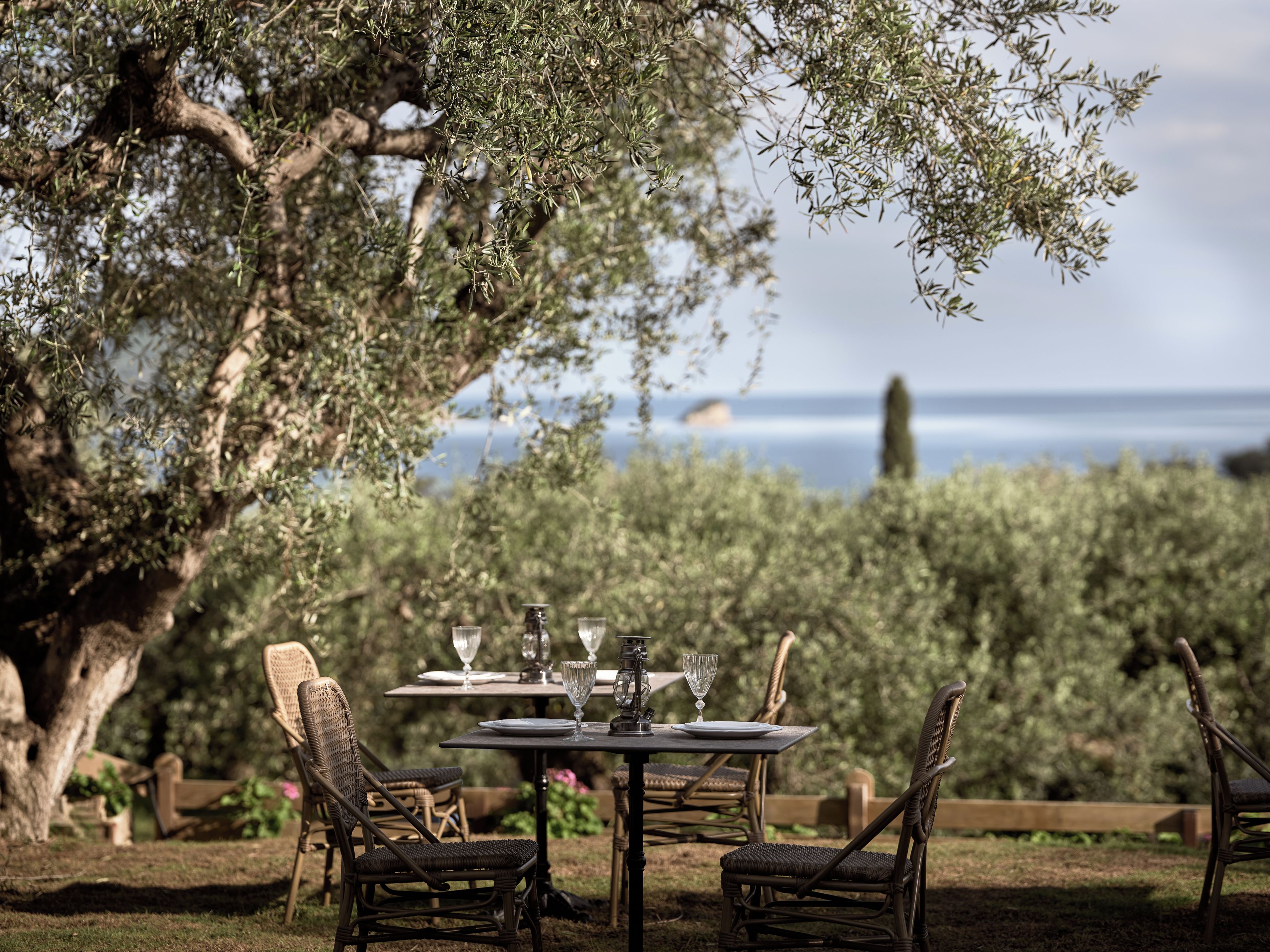 An outdoor dining area with wicker chairs and a table set for two, overlooking a garden and a view of the sea.