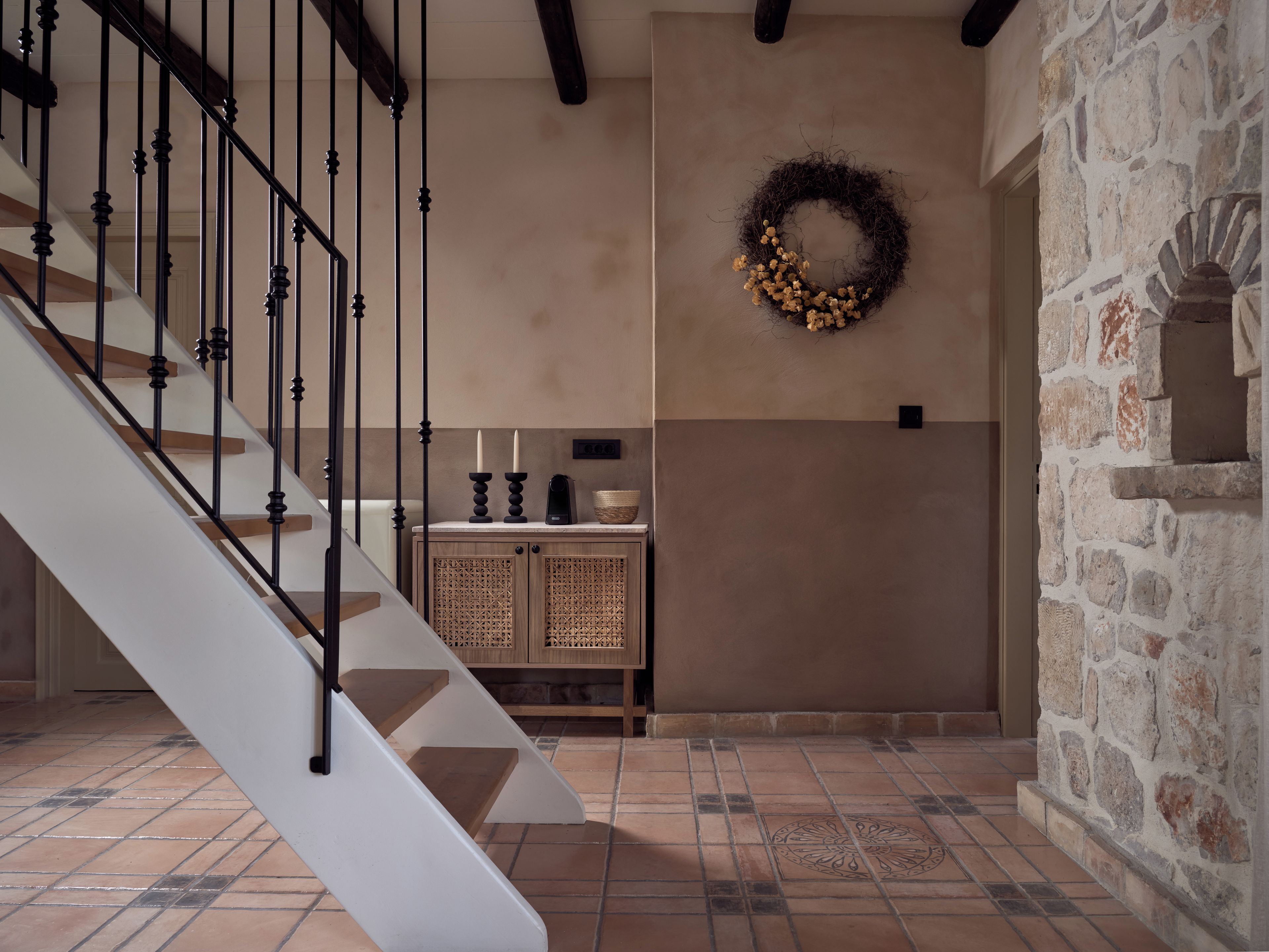 Una escalera con barandillas negras, una pared con una cesta tejida y velas, y una pared de piedra con un nicho decorativo.