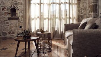 A cozy room with a stone wall, a large window with sheer curtains, a wicker side table, a round wicker stool, and a cushioned sofa.
